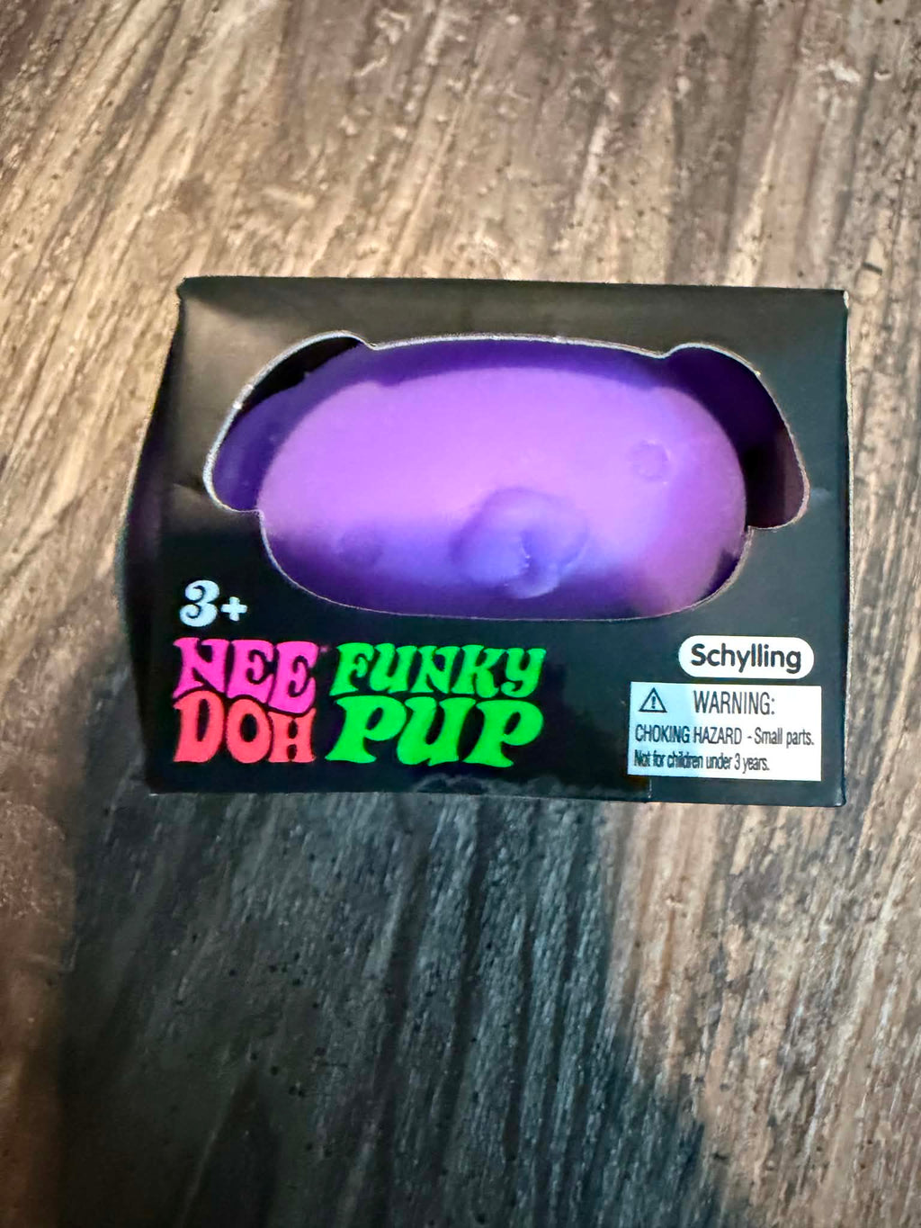 A boxed novelty squishy toy labeled 'Nee Doh Funky Pup' in purple, shown in its black retail packaging on a wood floor. The brand name 'Schylling' is visible on the box.