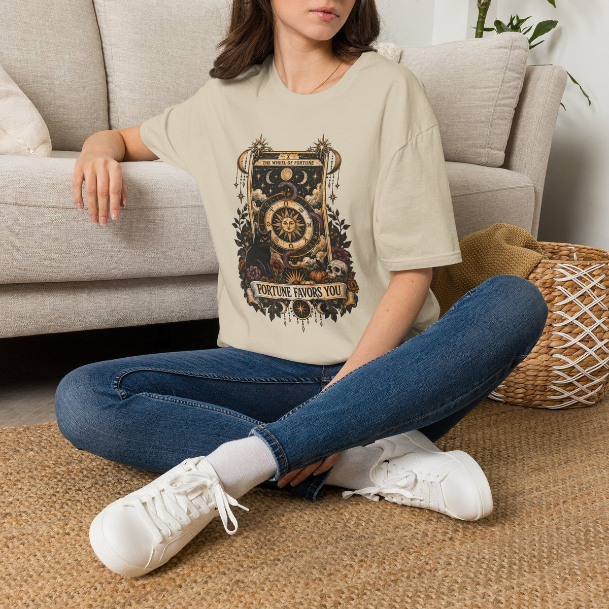 Woman sitting cross legged on a floor wearing a tan colored shirt with tarot card and cat that reads fortune favors you. 