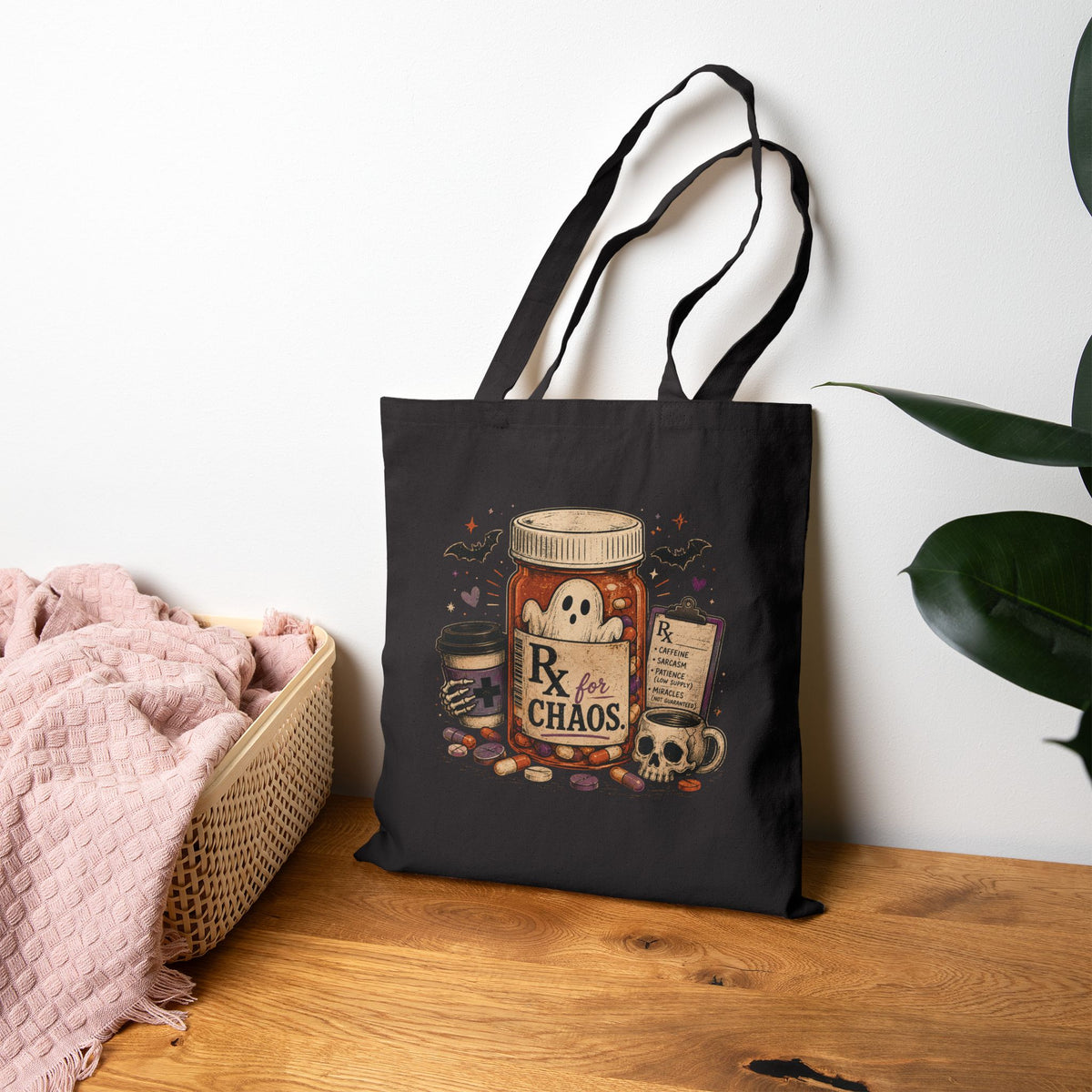 A black canvas tote bag with a Halloween-themed graphic print featuring a jar of pills labeled 'Rx for CHAOS', a skull, a ghost, and a potion bottle, displayed on a wooden table next to a pink blanket and a green plant.