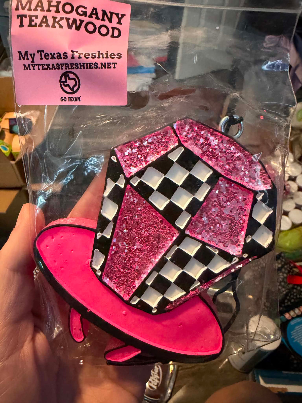 A hand holds a pink and black novelty teapot-shaped keychain inside a clear plastic bag with a pink 'MAHOGANY TEAKWOOD' sticker. The keychain features a black and white checkered pattern on the upper portion with glittery pink triangular panels, and a solid hot pink lower portion.