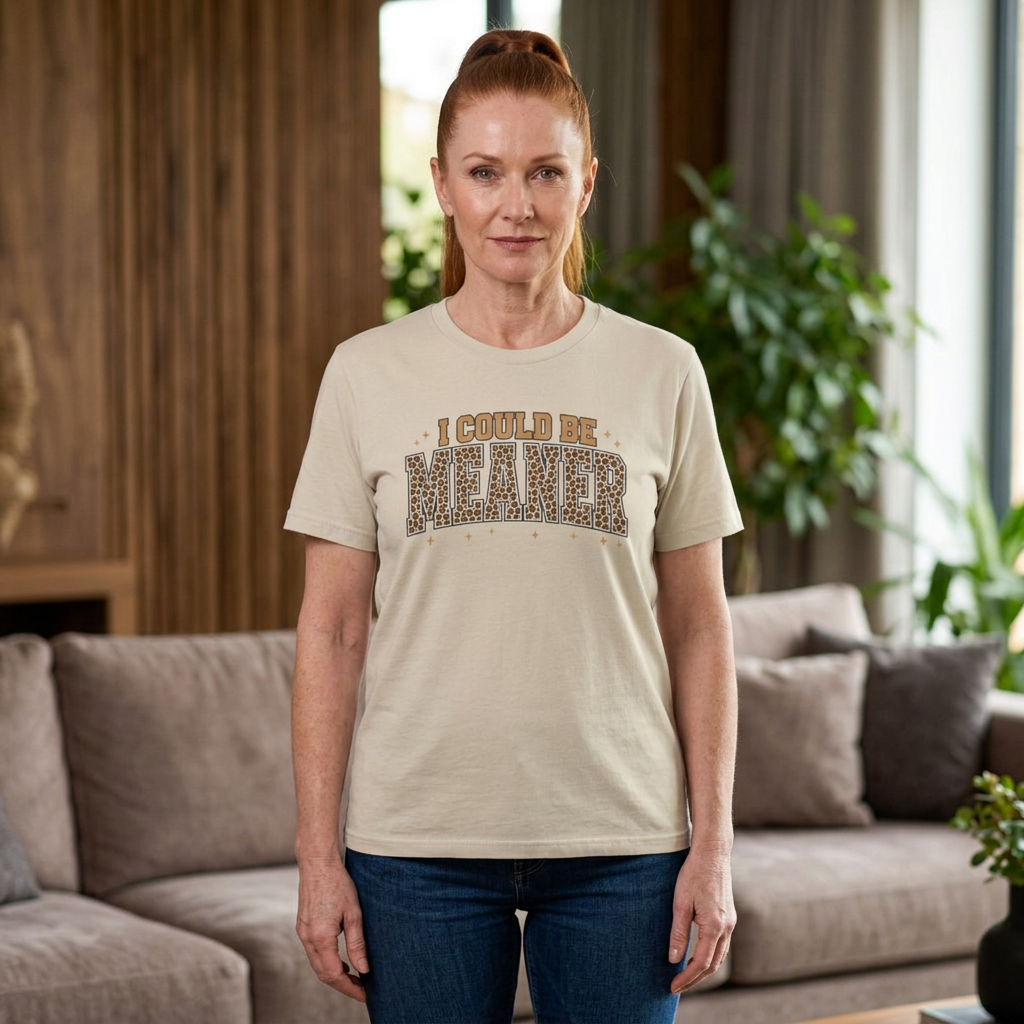 A woman wearing a beige graphic t-shirt with the text 'I COULD BE MEANER' in leopard print, standing in a living room with a couch and plants in the background.