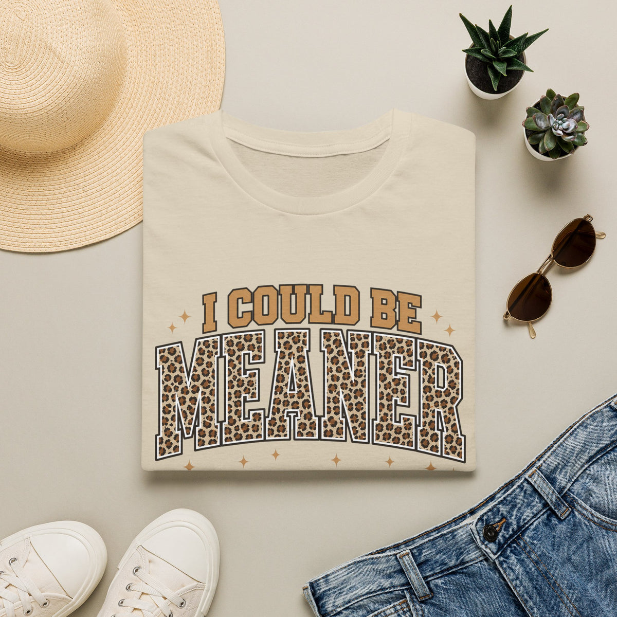 A folded cream graphic T-shirt is styled in a flat lay with a straw hat, white sneakers, blue jeans, sunglasses, and small potted succulents. The shirt reads 'I COULD BE MEANER' with the word 'MEANER' in a leopard-print pattern.