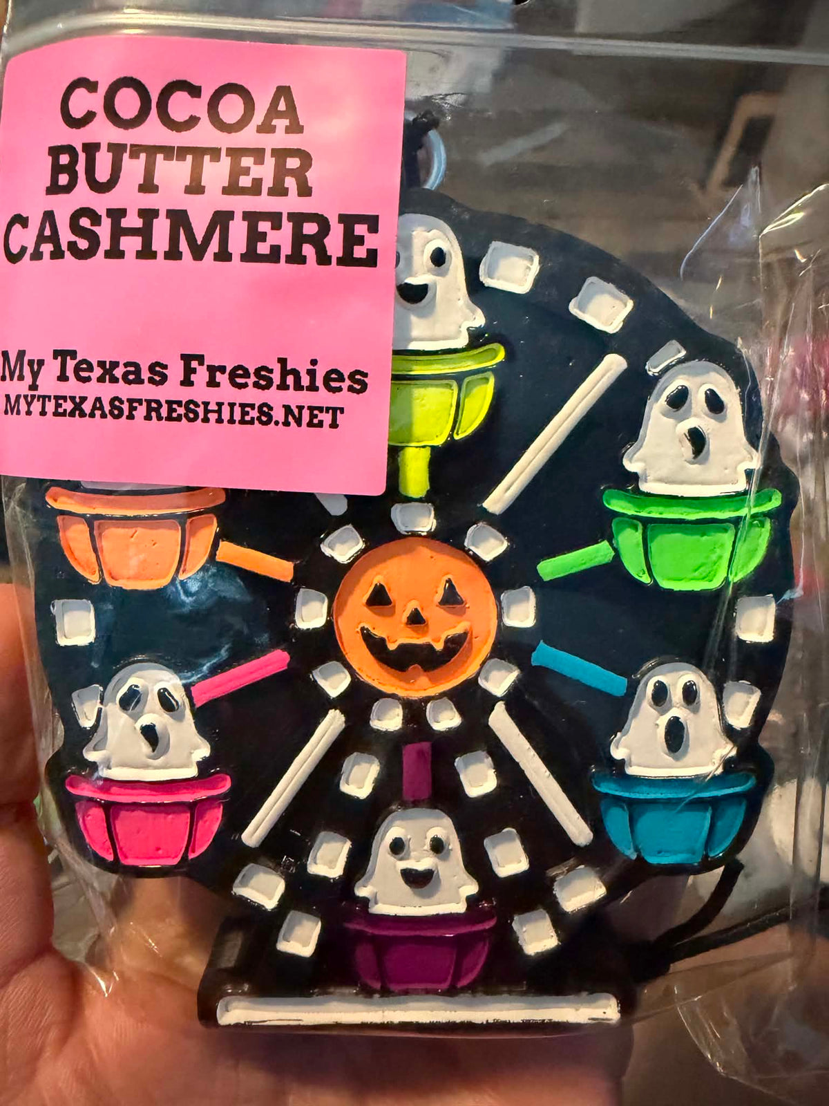 A hand holding a packaged Halloween-themed cookie shaped like a Ferris wheel. The cookie has ghost cupcake cars in orange, green, pink, blue, and purple, a central orange jack-o'-lantern, and white accents on a dark base.