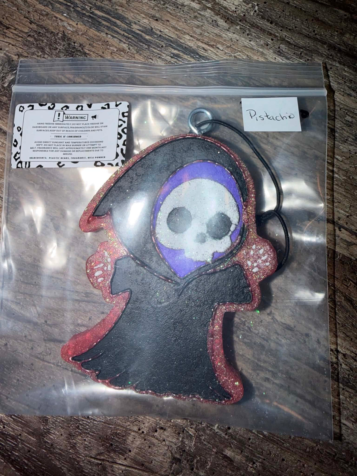 A handmade gothic-style grim reaper ornament inside a clear plastic bag, featuring a black felt robe with red glitter trim, a purple glitter halo, and a white skull face. A small handwritten note says 'Pistache' and a brand card is visible on the left.