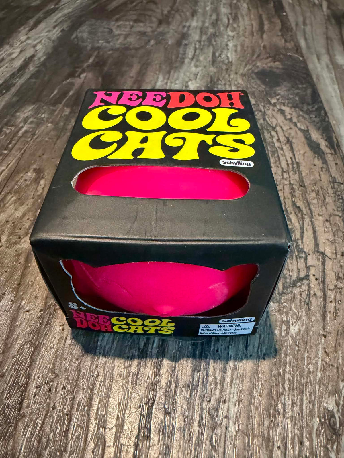 A black cardboard box for the 'Needoh Cool Cats' toy, featuring a bright pink cat-shaped ball visible through a cutout window on the front and side. The box has bold retro-style yellow and pink lettering on a dark background and is photographed on a dark wood floor.