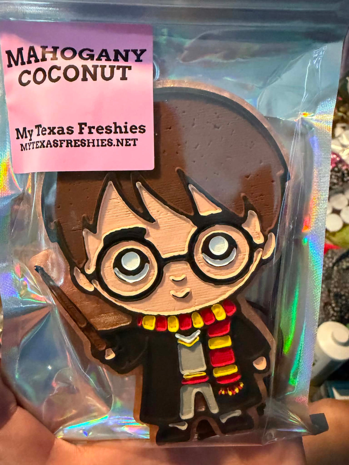 A packaged novelty cookie shaped like a cartoon Harry Potter character with glasses, a black robe, and a red-and-yellow striped scarf, sealed in a clear plastic bag with a pink label reading 'Mahogany Coconut' and 'MyTexasFreshies'.
