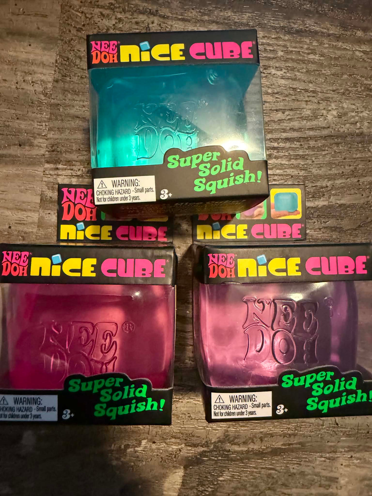 Four boxed 'Super Solid Squishy!' Nee Dot Nee Do products arranged on a wood surface. The packages show translucent jelly-like cubes in blue-green, pink, and magenta, with visible 'Nee Dot Nee Do' branding and warning labels.