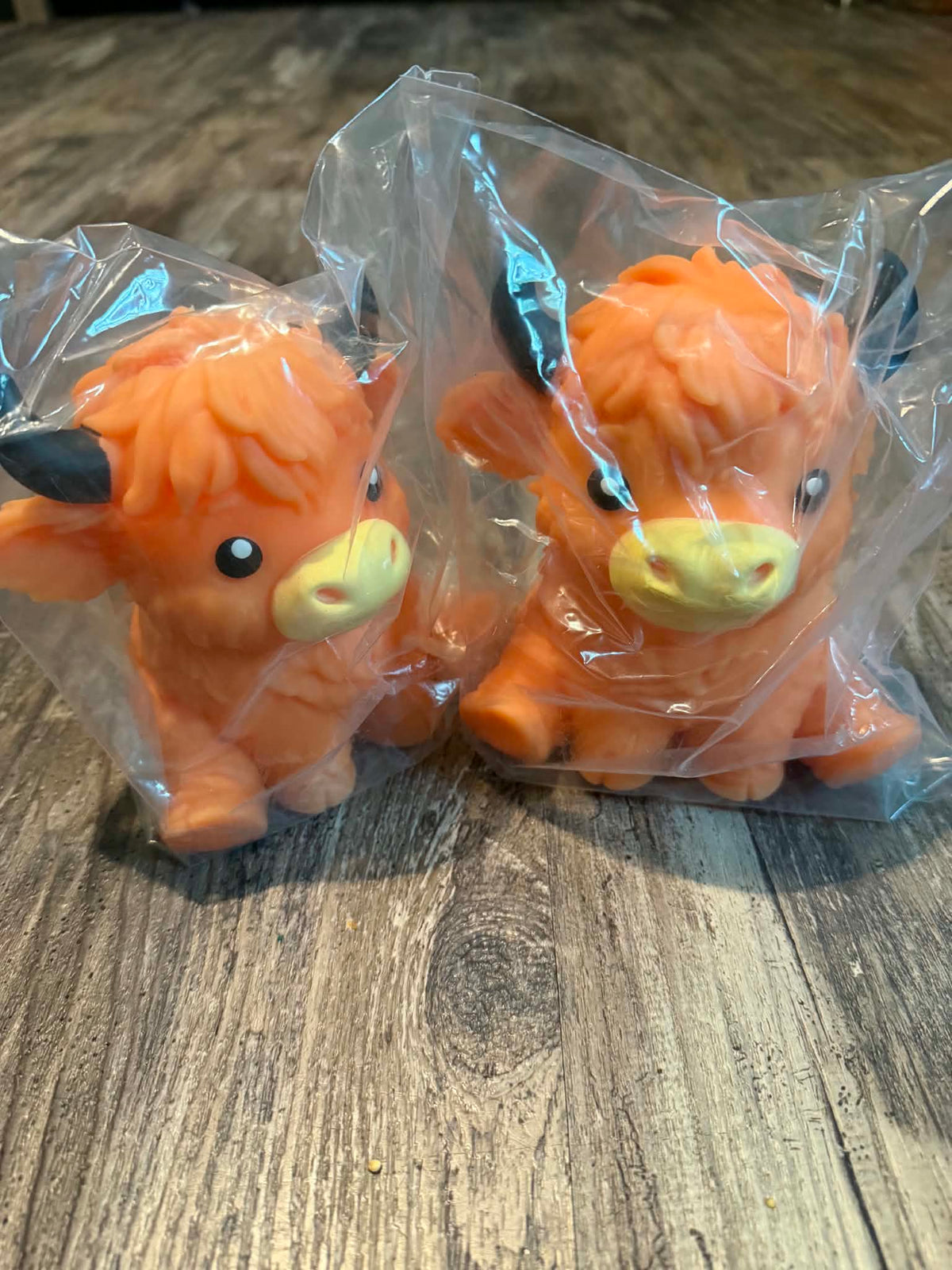 Two small orange cow figurines in clear plastic packaging, placed on a wooden surface.