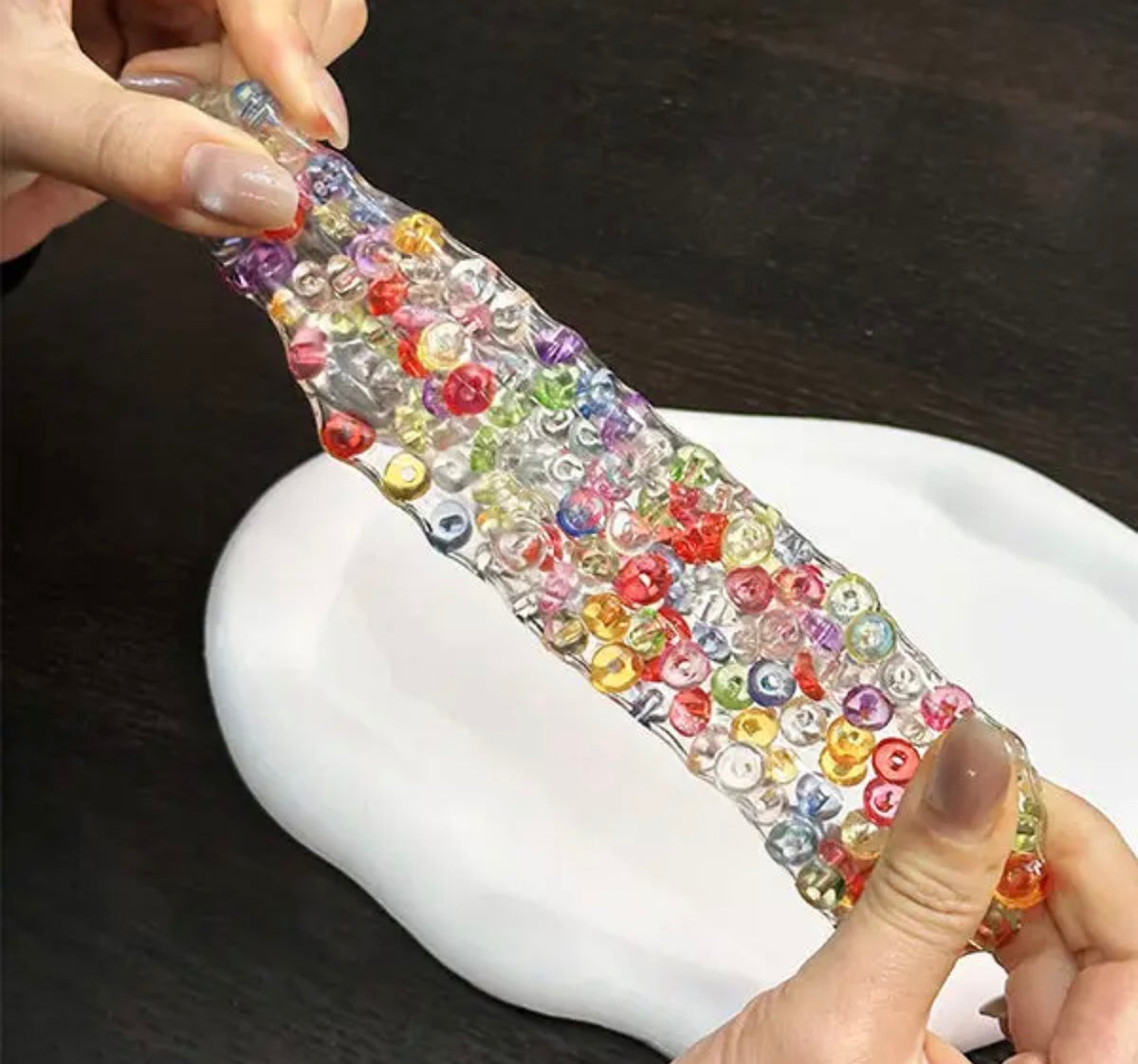 A person stretches a translucent, elastic slime-like strip filled with many small colorful round beads over a white tray on a dark tabletop.