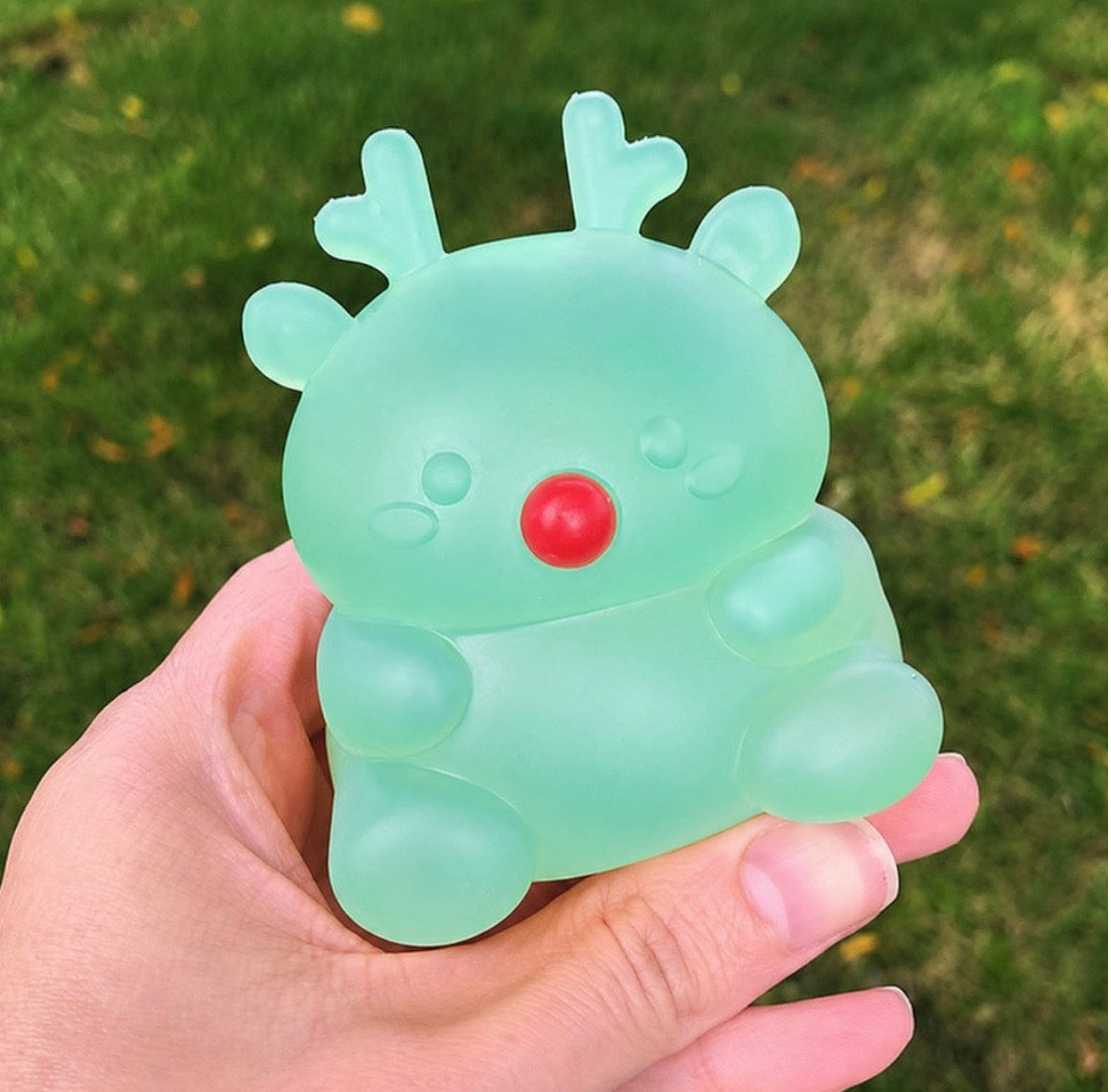 A hand holding a translucent mint green reindeer-shaped object with a red nose against a blurred green grass background.