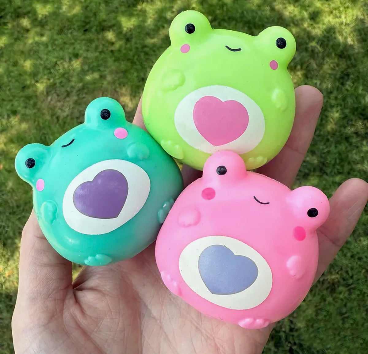 Three cute frog-shaped squishy stress toys in green, teal, and pink, held in a person's hand against a blurred green grass background.