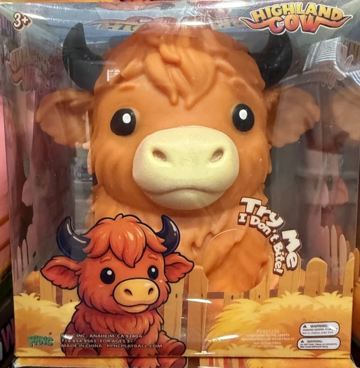 Highland Cow toy in its original retail packaging, featuring a large orange-brown plush cow with black horns and a cream-colored snout, alongside a smaller cartoon Highland Cow character on the front of the box.