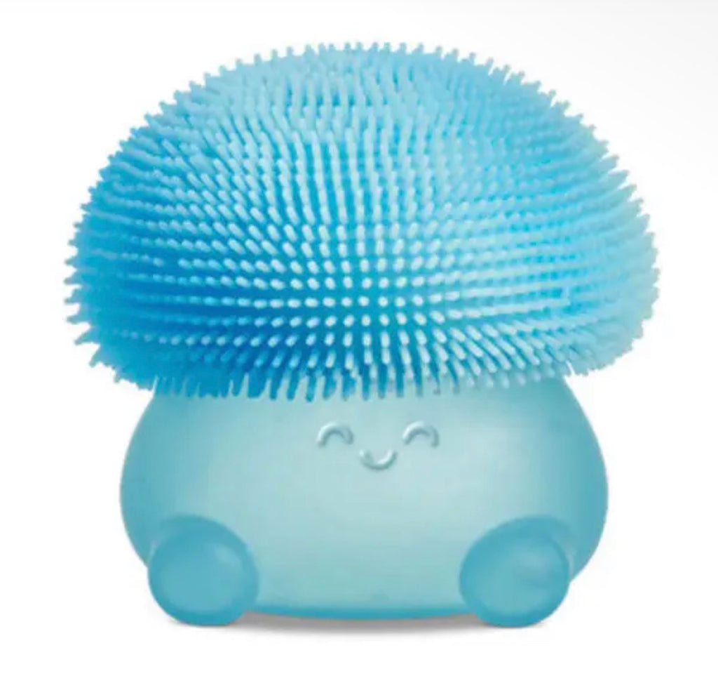 A light blue silicone facial cleansing brush with a rounded base, small feet, and a large, dome-shaped bristle head. The base features a simple embossed smiley face.
