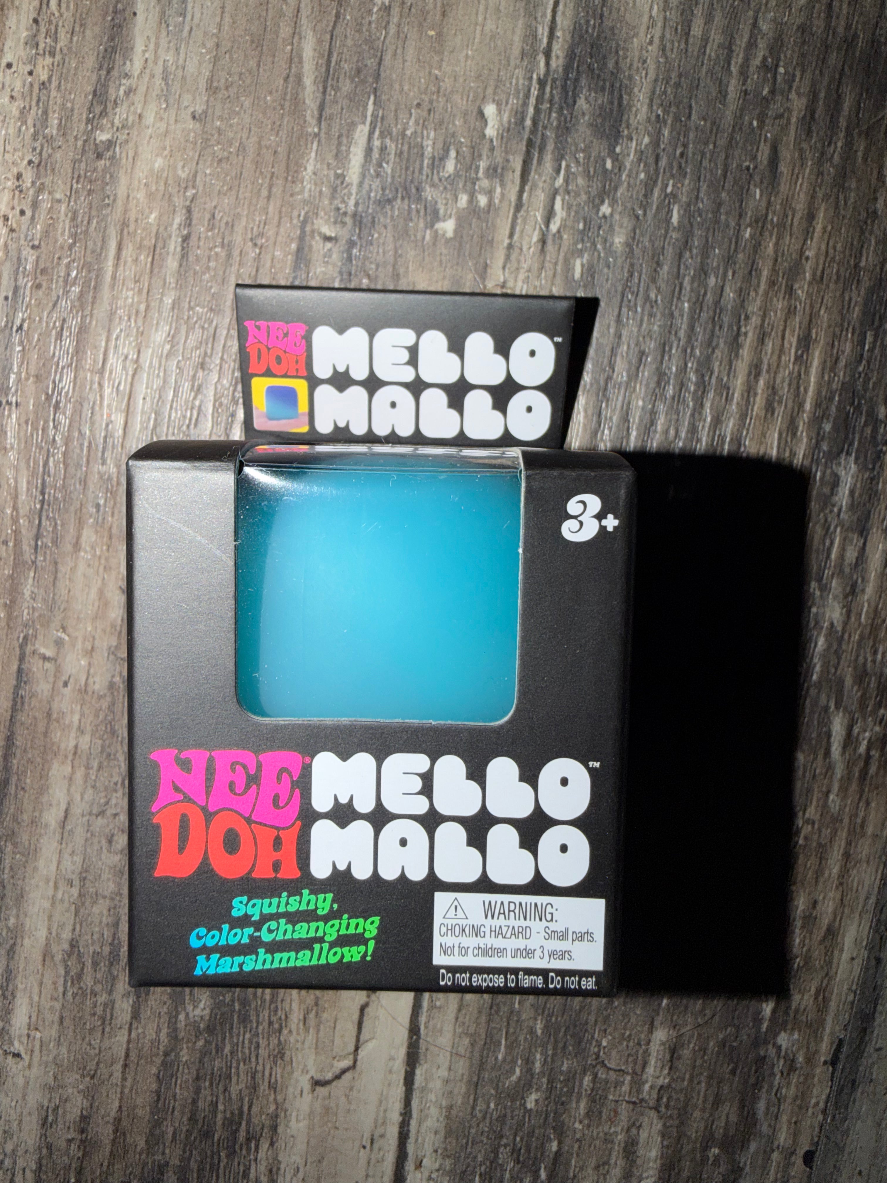 A package of Nee Doh Mello Mallo squishy, color-changing marshmallow toy in a bright turquoise blue color, shown in its original black retail box on a dark wood background.