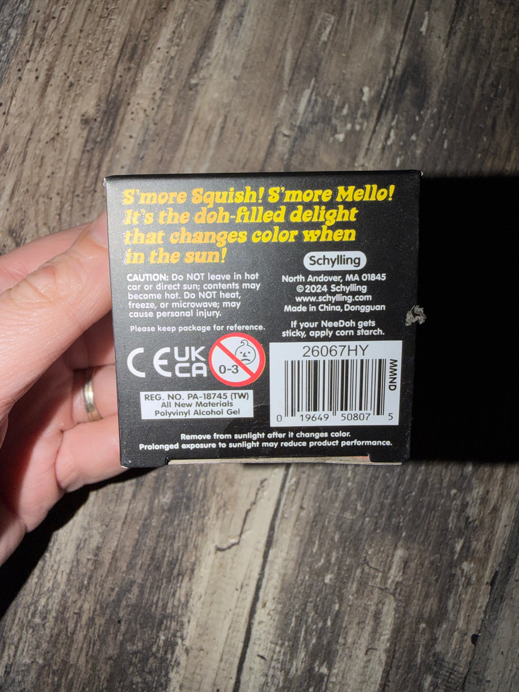 A hand holding a black box of Schilling S'more Squish! S'more Mello! against a weathered gray wood background. The back of the box features yellow and white text, warning labels, and a barcode.