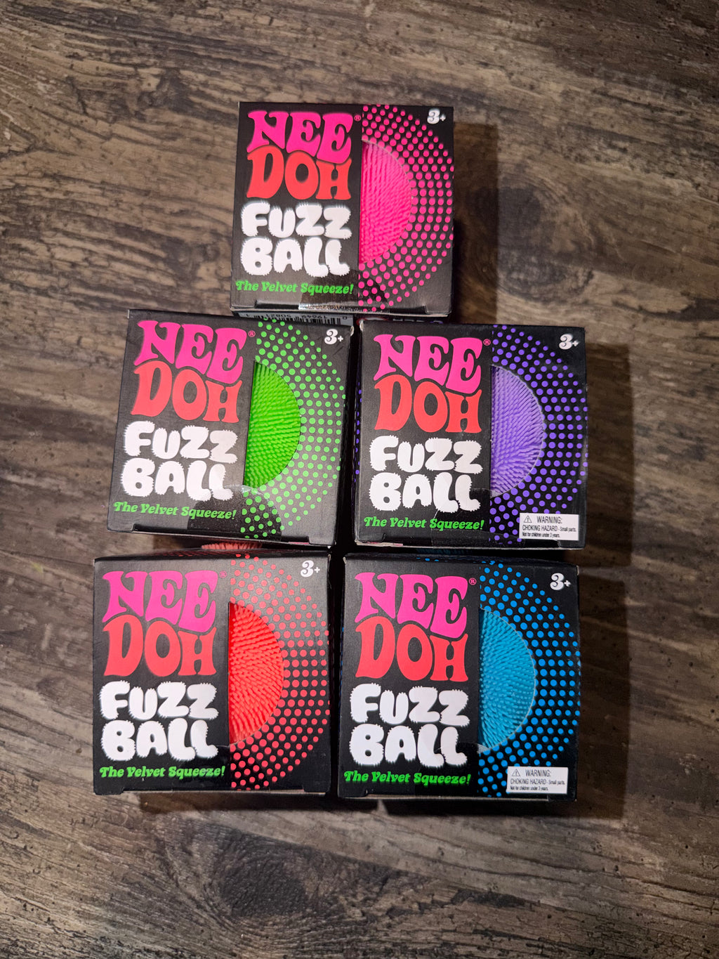 Six boxes of Nee Doh Fuzz Ball pool balls arranged in two rows on a dark wood surface. The boxes feature different colored fuzz balls: pink, green, purple, red, and blue, with the brand name 'NEE DOH' and product name 'FUZZ BALL' clearly visible.