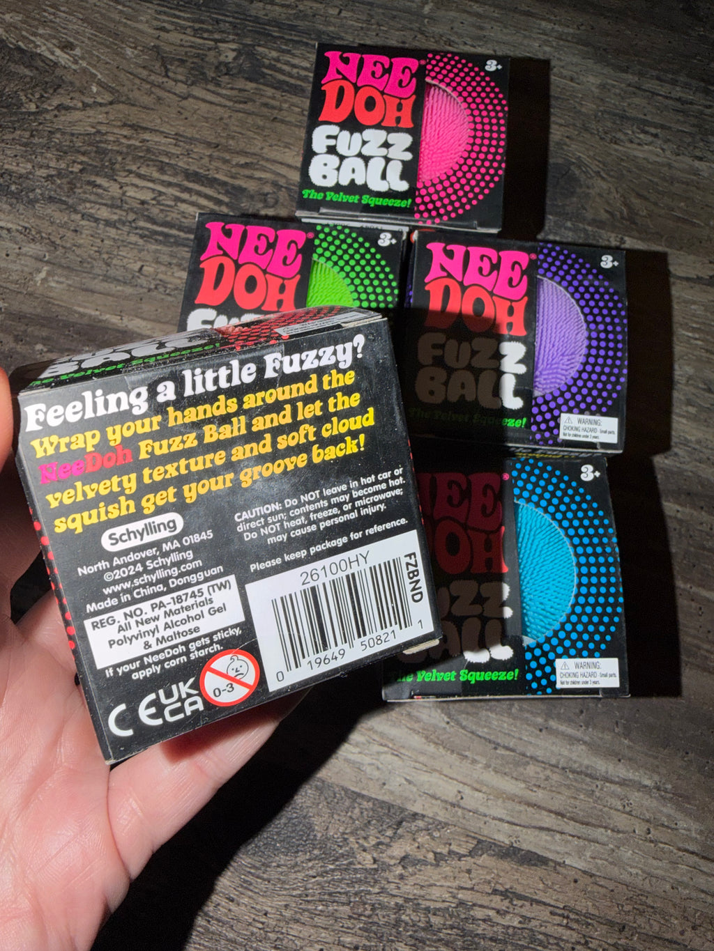 A hand holds a box of NeeDoh Fuzz Ball adult novelty product, with several other colored boxes stacked behind it on a dark wood surface.