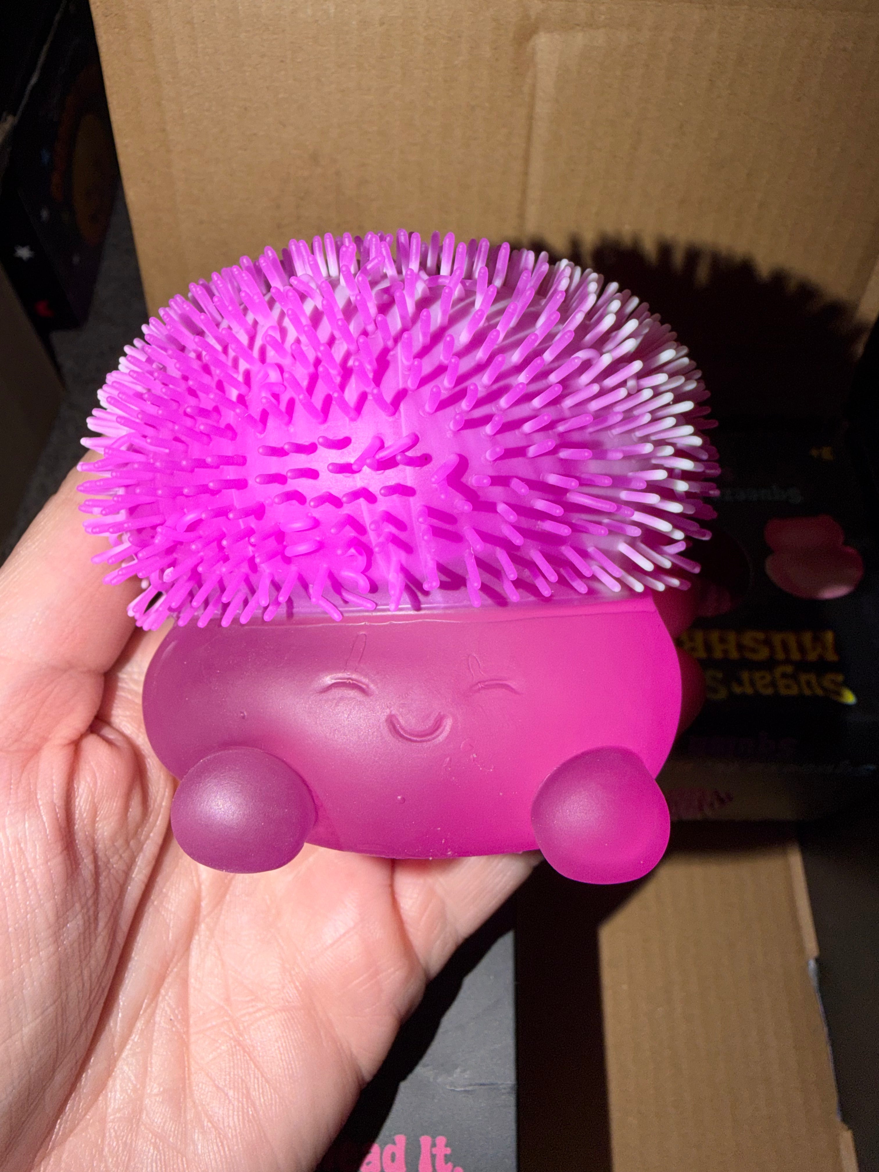 A hand holding a bright pink mushroom-shaped silicone scrubber with a spiky crown and smooth base, photographed indoors against a cardboard box background.