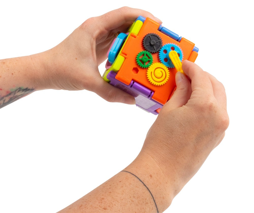 A close-up of a person's hands holding and operating a colorful plastic sensory toy with gears and levers, isolated on a white background.