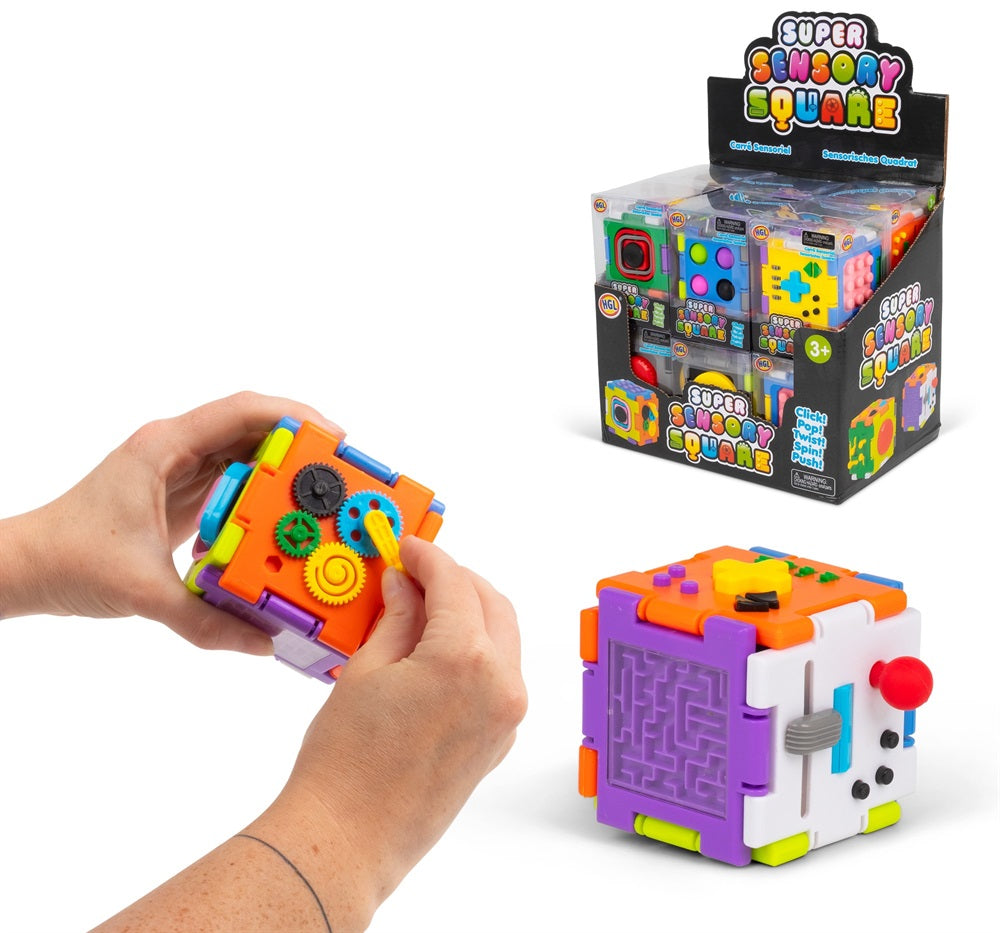 A colorful sensory cube toy shown being held by two hands, with one cube displayed on a white surface and its retail display box in the background. The toy features multiple interactive panels including dials, gears, a maze, and buttons in bright colors.