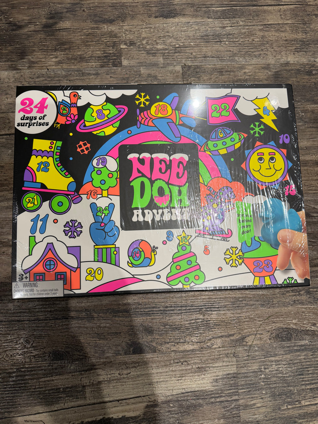 A colorful 'Nee Doh Advent' 24-day surprise box set on a dark wood floor, sealed in clear plastic wrap with a hand holding the bottom right corner.
