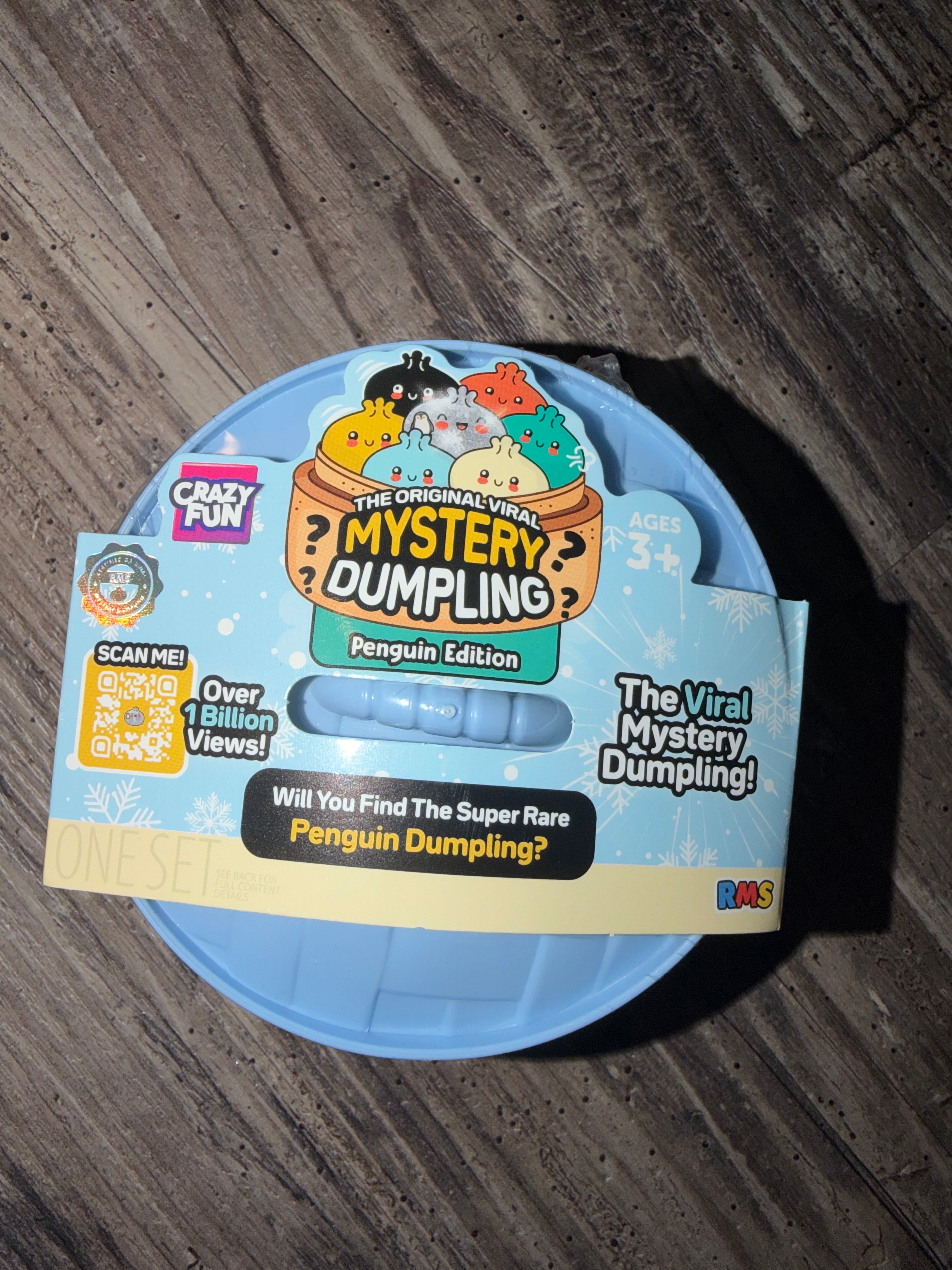 A light blue plastic mystery dumpling container with its retail packaging, placed on a dark wood floor. The packaging features cartoon penguin graphics and text for 'The Original Viral Mystery Dumpling Penguin Edition' by Crazy Fun.