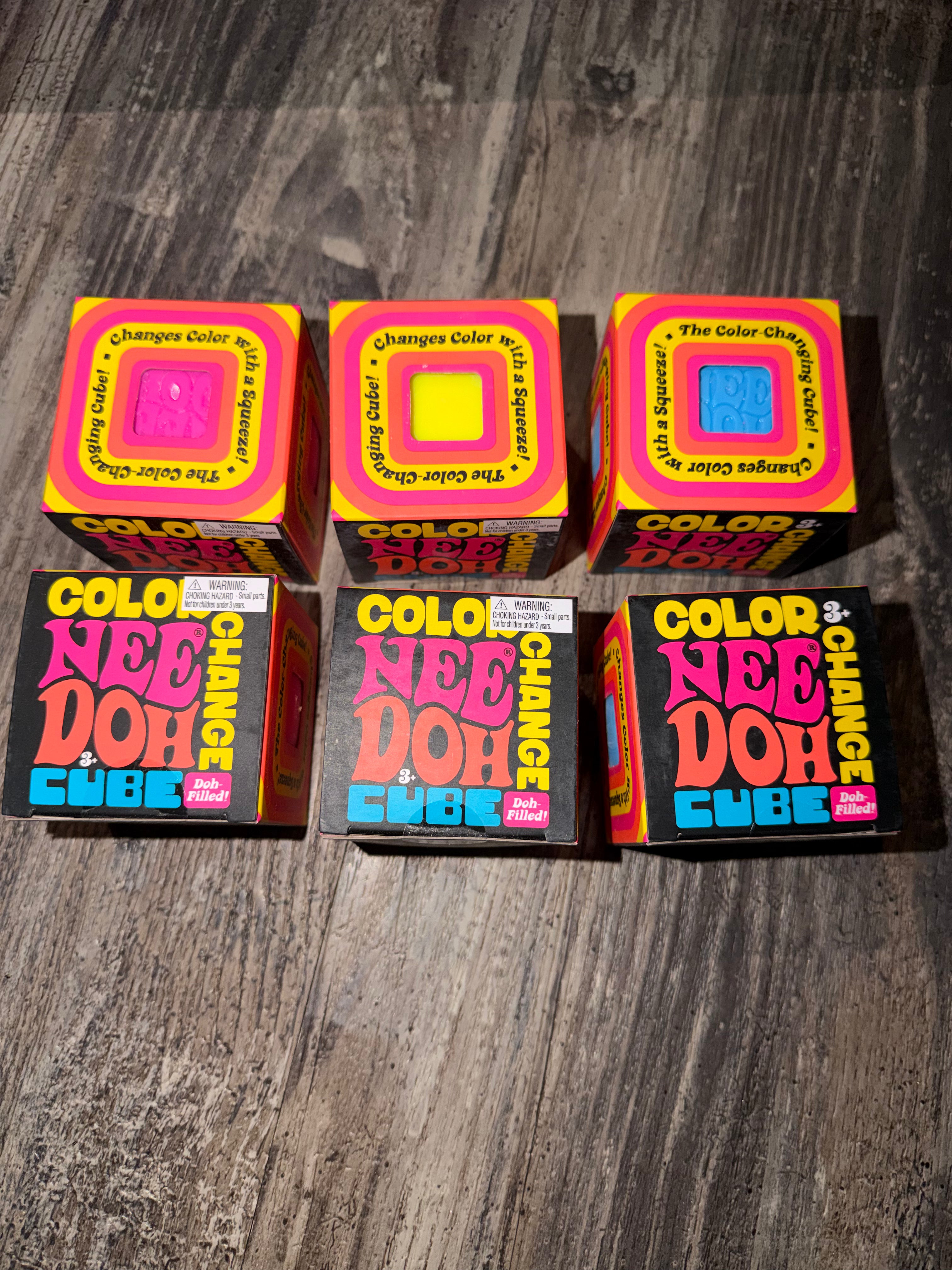 Six boxes of Color Nee Doh Change Color Cube toys arranged in two rows on a dark wood floor. The top row contains three boxes showing the colorful changing-dough face, and the bottom row contains three boxes showing the product branding.