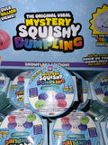 A retail display of Toy Biz 'The Original Viral Mystery Squishy Dumpling' Snowflake Edition packages, showing multiple round plastic containers with colorful printed labels in front of a larger blue promotional sign.