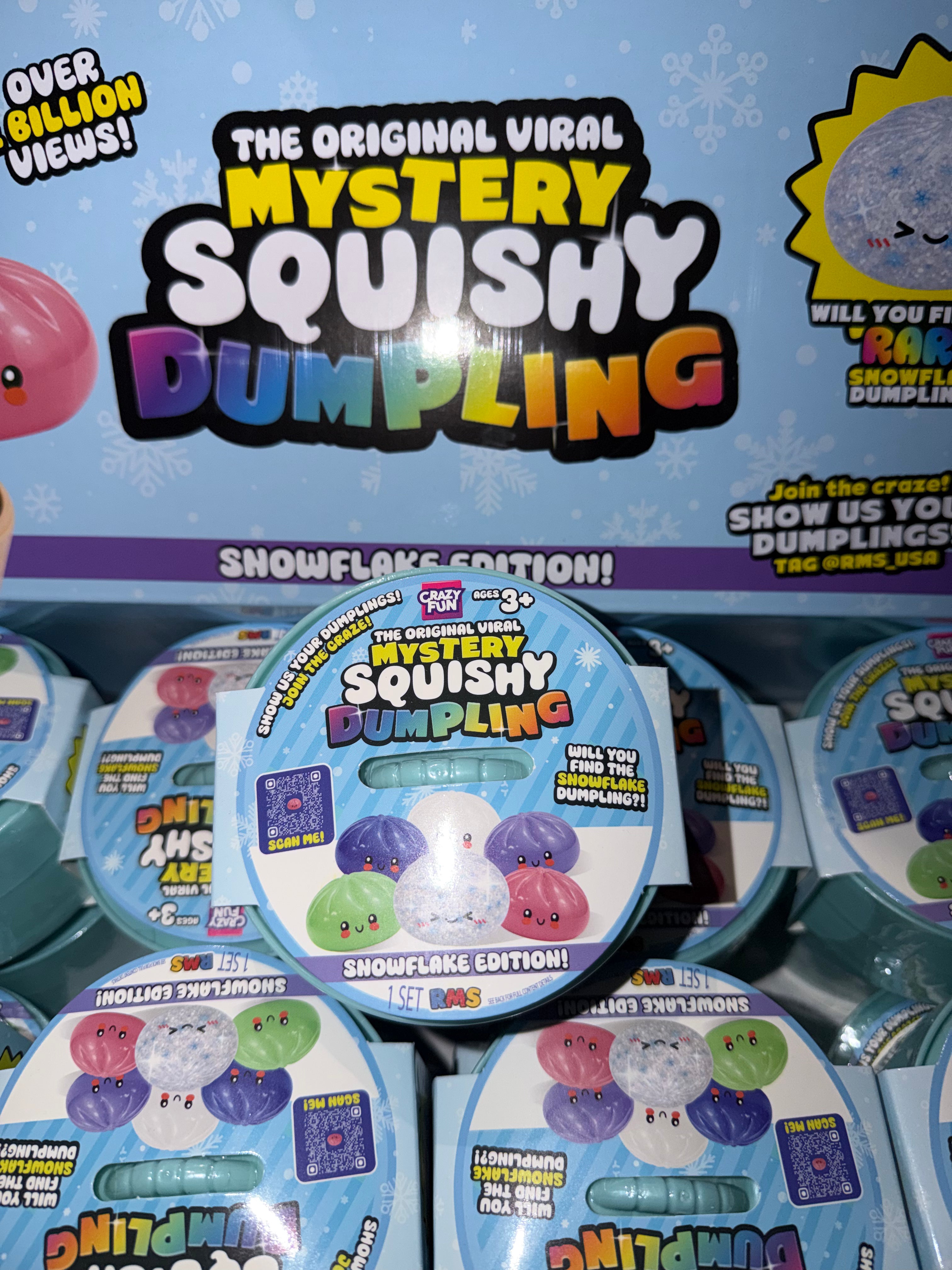 A retail display of Toy Biz 'The Original Viral Mystery Squishy Dumpling' Snowflake Edition packages, showing multiple round plastic containers with colorful printed labels in front of a larger blue promotional sign.