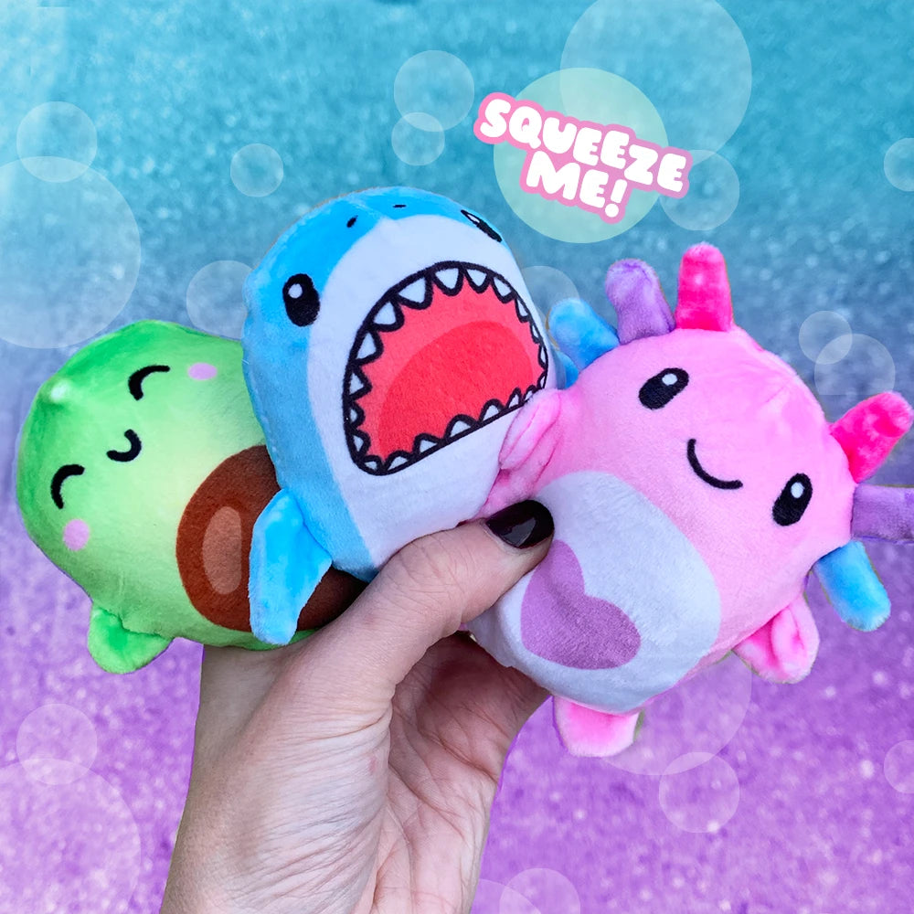 A hand holds three plush shark-themed squishy toys against a colorful blue and purple background with bubble graphics. The toys include a green avocado character, a blue shark with an open mouth showing red teeth, and a pink axolotl-like shark with blue fins and a purple heart.
