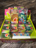 A display box filled with packaged novelty squishy toys and gummy-like items. The front packages read 'CRYSTAL SQUISHY' and 'Super Solid Squishy,' with colorful fruit-themed boxes arranged in a bright lime-green tray on a wood surface.