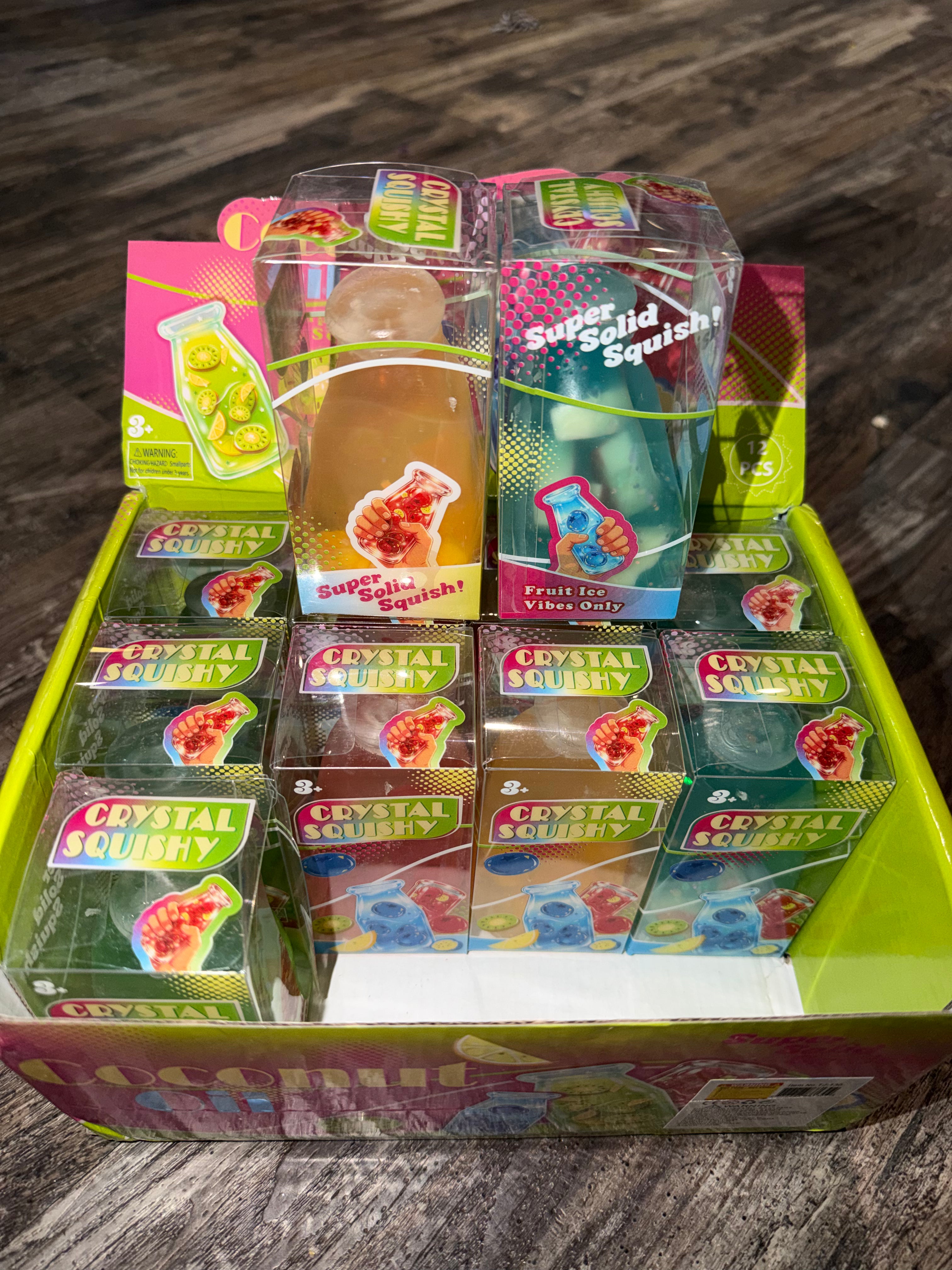 A display box filled with packaged novelty squishy toys and gummy-like items. The front packages read 'CRYSTAL SQUISHY' and 'Super Solid Squishy,' with colorful fruit-themed boxes arranged in a bright lime-green tray on a wood surface.