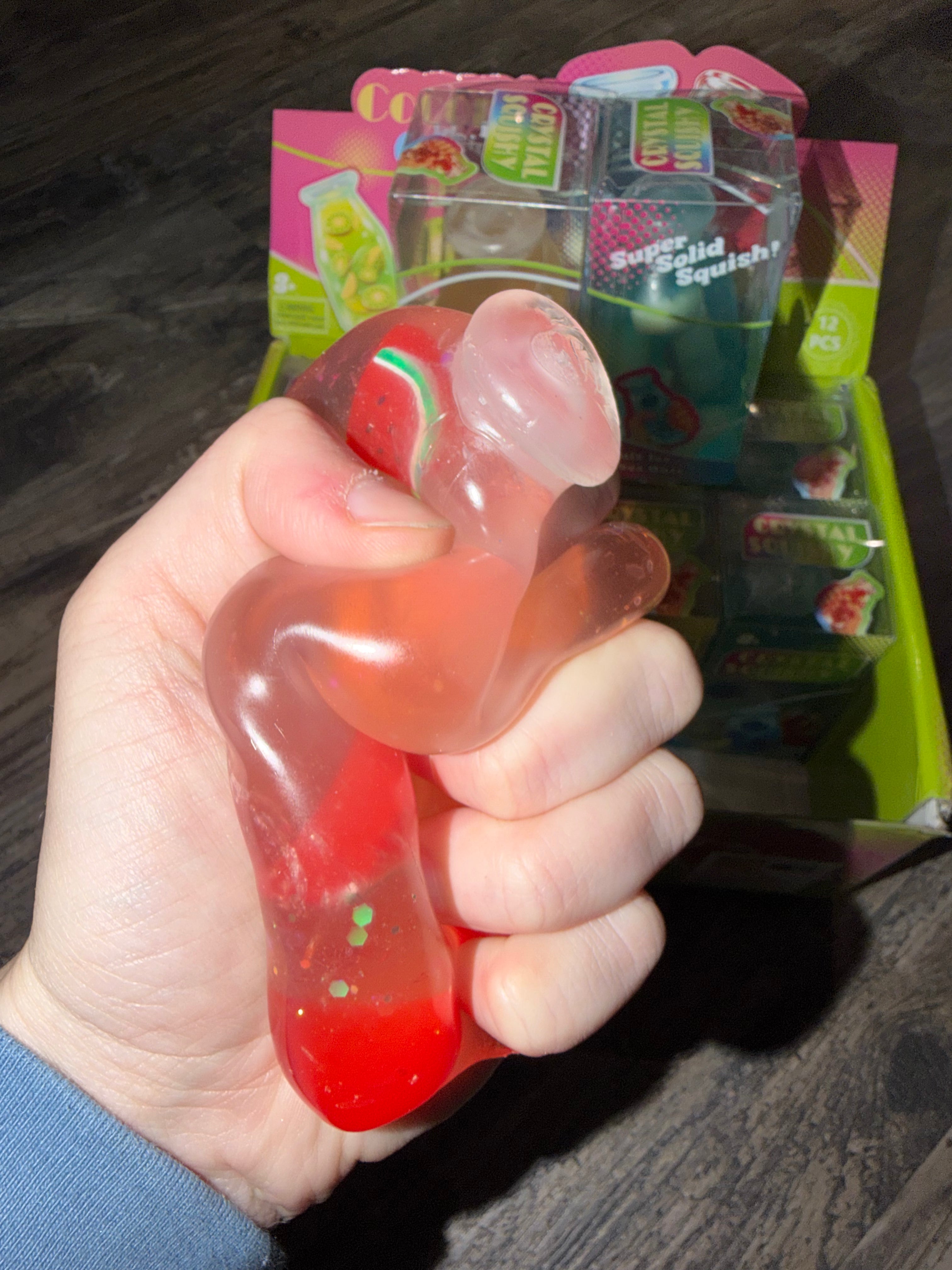 A hand holding a translucent red and pink squishy toy with green speckles, with a display box of similar toys in the background on a dark wooden surface.