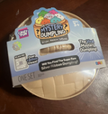 A packaged toy product labeled 'The Original Viral Mystery Dumpling' in the Silver, Ribbon Edition, shown on a dark wooden surface. The package contains a beige woven dumpling visible through a clear window, with colorful branding and product text on the top.