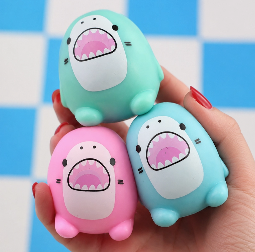 Three cute shark-shaped squishy stress toys in mint green, light blue, and pink, held in a hand against a blue and white checkered background.