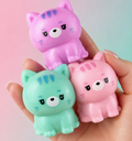 Three small cat-shaped squishy toys in pink, mint green, and light pink are being held in a person's hand against a soft pink-to-blue gradient background.