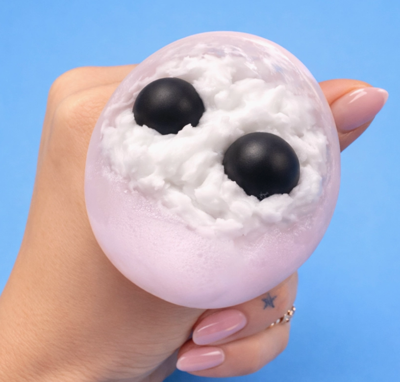 A hand with pink manicured nails holds a squishy, round novelty toy shaped like a white fluffy cloud or foam ball with two black oval eyes, against a solid blue background.