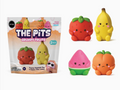 A 2-pack of 'The Pits Grumpy Fruits' foam squeeze toys in their packaging, shown alongside the five individual fruit-shaped toys: a strawberry, a banana, a watermelon slice, an orange, and a panda. The toys are made of colorful, squishy foam with cute grumpy faces.