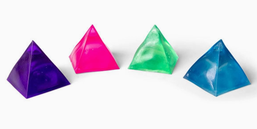 Four translucent pyramid-shaped gelatin or jelly cubes in purple, pink, green, and blue, arranged in a row on a plain white background.