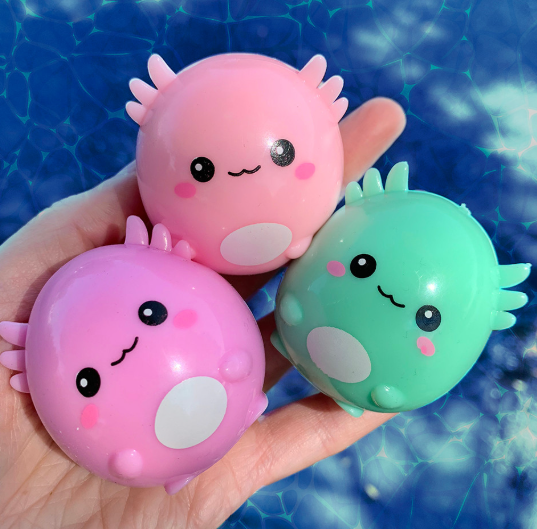 Three small, round, axolotl-shaped novelty items in pink and mint green are held in a person's hand against a blue water background.