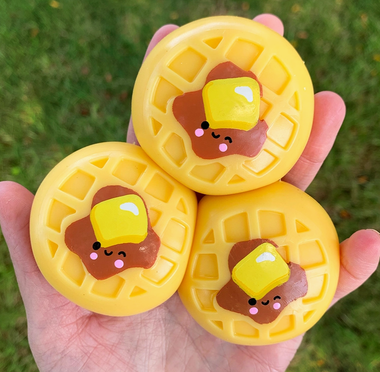 Three yellow waffle-shaped squishies with cute butter and bacon faces are held in a person's hand against a blurred green grass background.