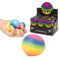 A hand squeezes a rainbow-colored stress ball, with a larger rainbow ball in the foreground and a display box of 'Rainbow Squishy Balls' behind it on a white background.