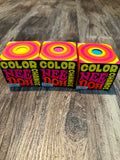 Three colorful boxes of 'Color Change Nee Doh' toys arranged side-by-side on a rustic wooden surface. The packaging features a retro psychedelic design with concentric circles in yellow, pink, and orange, and bold text reading 'COLOR CHANGE' and 'NEE DOH'.
