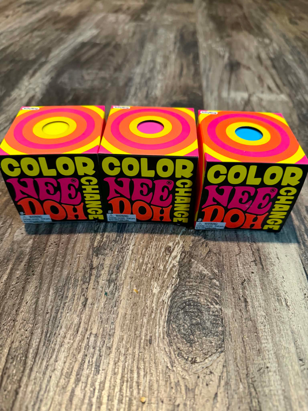 Three colorful boxes of 'Color Change Nee Doh' toys arranged side-by-side on a rustic wooden surface. The packaging features a retro psychedelic design with concentric circles in yellow, pink, and orange, and bold text reading 'COLOR CHANGE' and 'NEE DOH'.