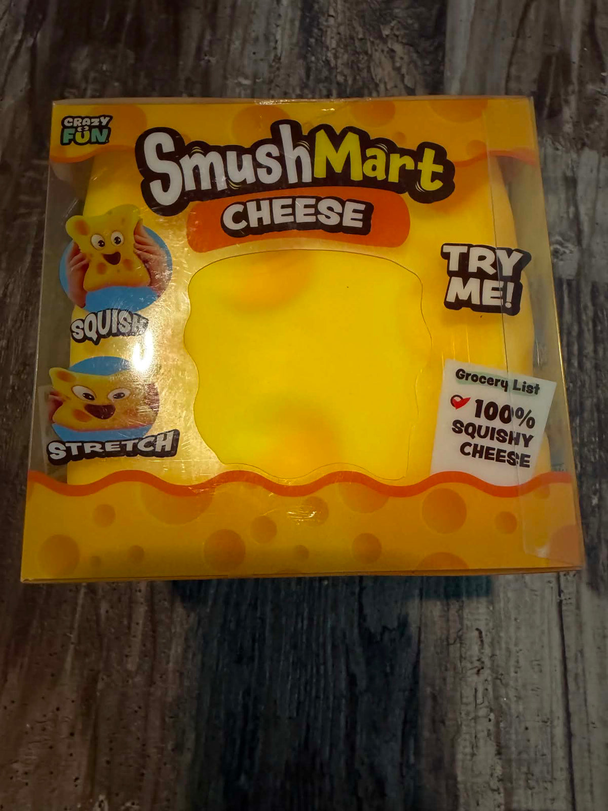 SmushMart Cheese squishy toy in its original packaging, featuring a yellow cheese-shaped squishy inside a clear plastic box with cartoon graphics and text on a dark wood background.