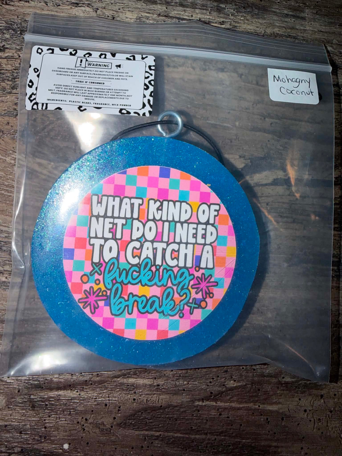 A round glittery blue and pink decorative sign with the text 'WHAT KIND OF NET DO I NEED TO CATCH A fucking break?' inside a clear plastic bag on a wooden surface.