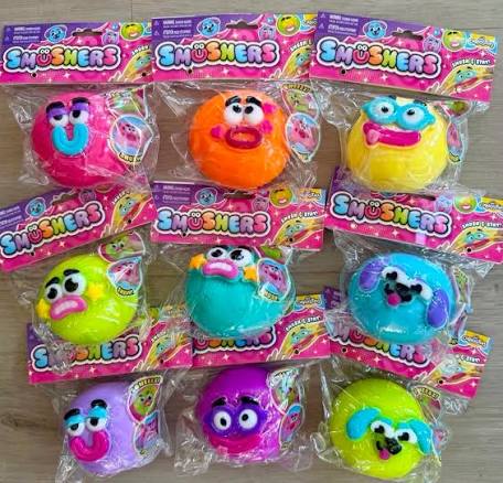 Nine packaged Smushers squishy toys in various bright colors, arranged in a grid on a light wood surface. Each toy is in its original blister packaging with a pink and purple header card.